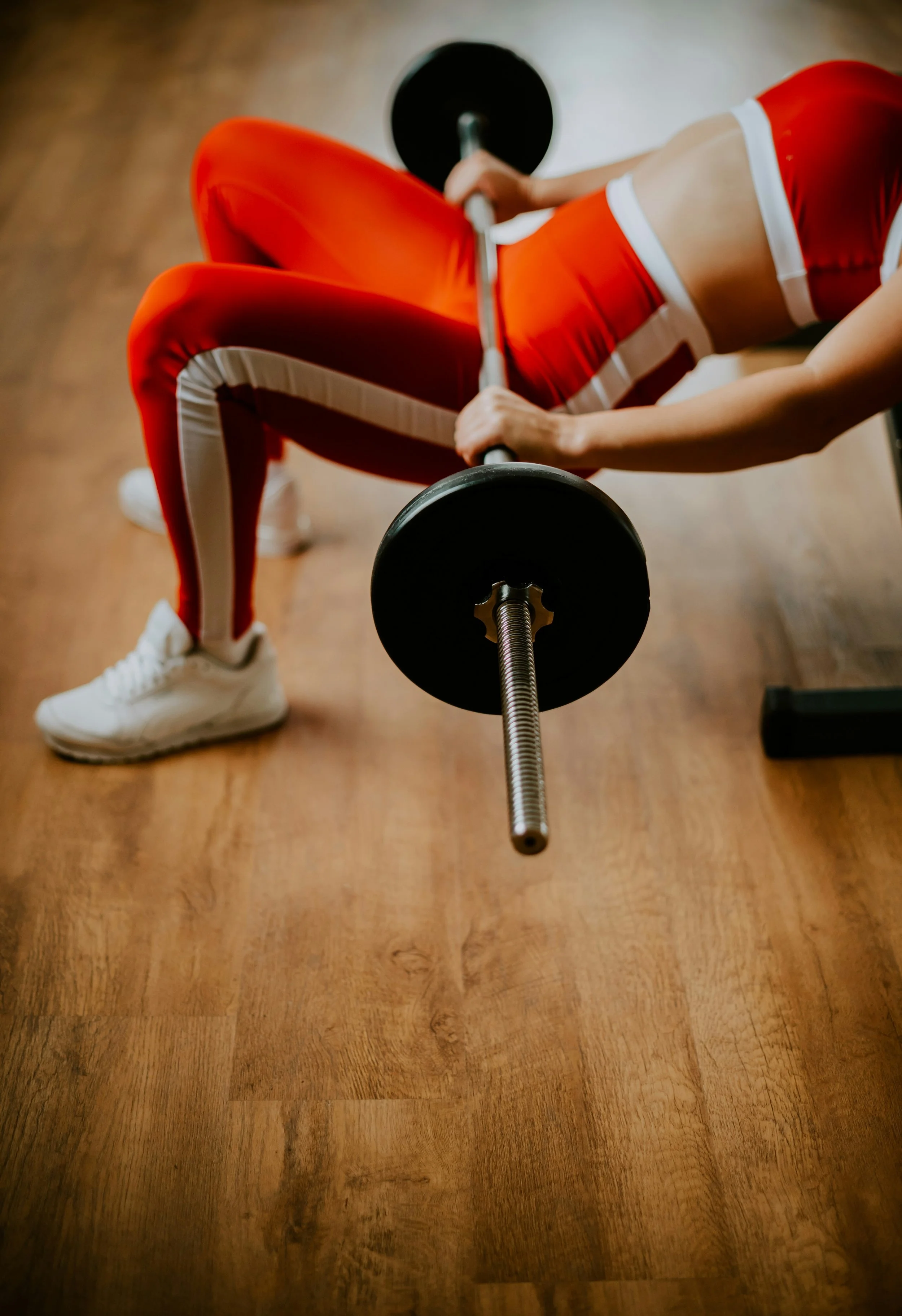 A woman in athletic clothing performing a bench press exercise with a barbell on a wooden floor gym.