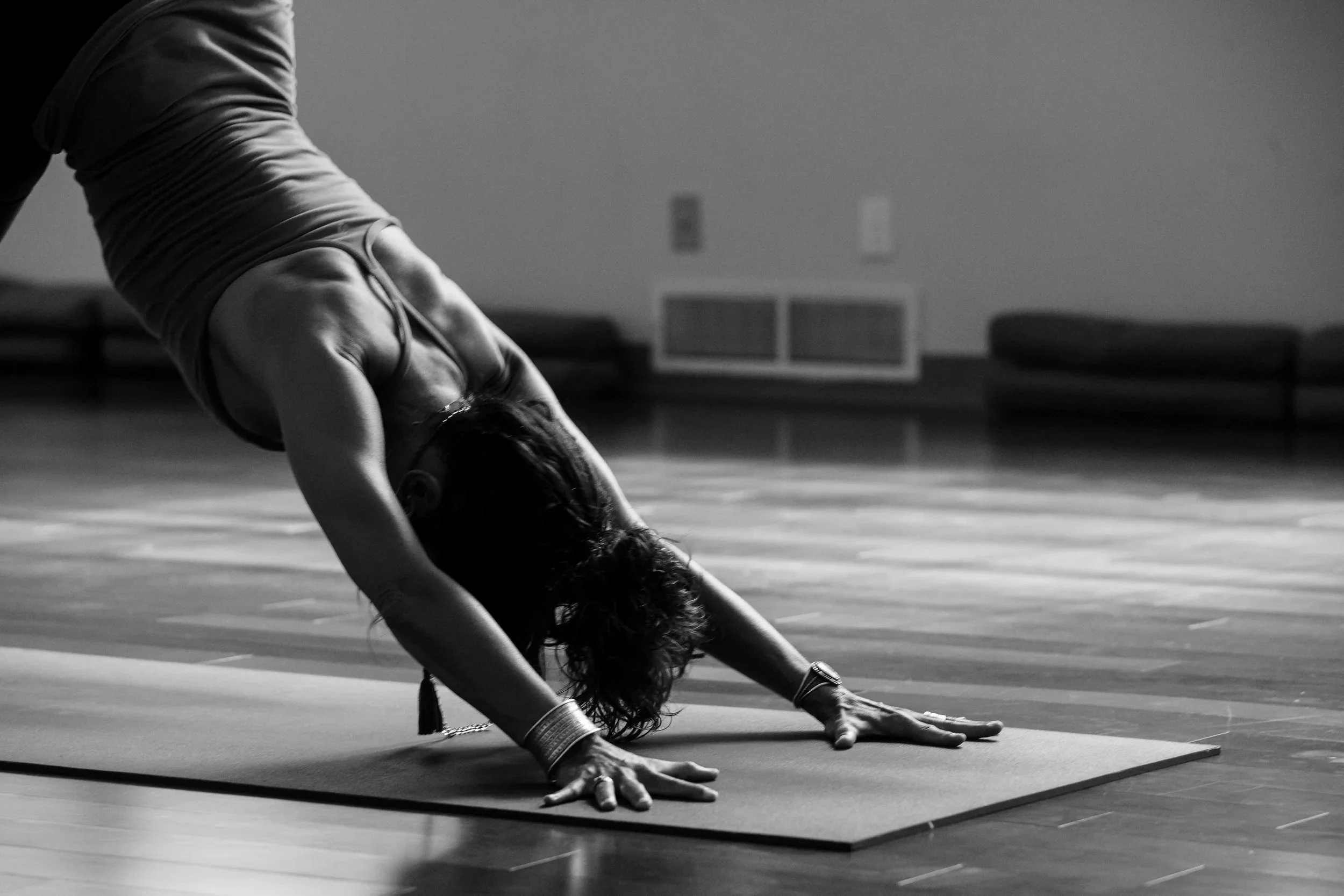 A person in workout attire performing a downward dog yoga pose on a mat in a room with wooden flooring, minimal furniture, and plain walls.