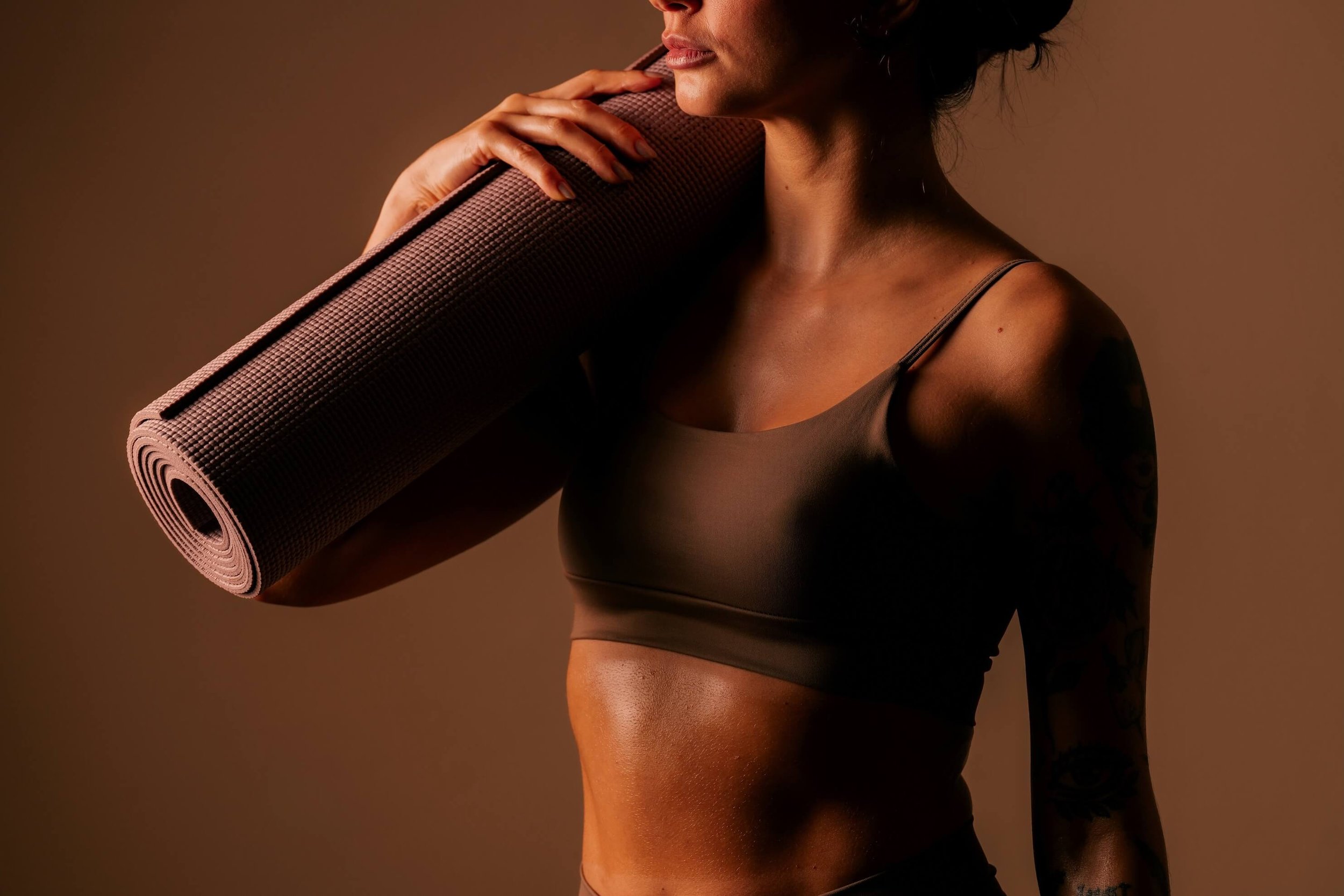 A woman in athletic attire holding a rolled yoga mat over her shoulder, with part of her face and body visible against a warm brown background.