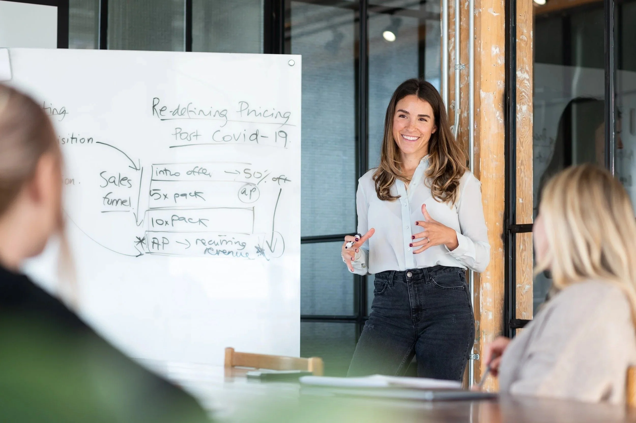 A woman giving a presentation or lecture in front of a whiteboard, smiling and gesturing with her hands, with an audience of several people seated in front of her.