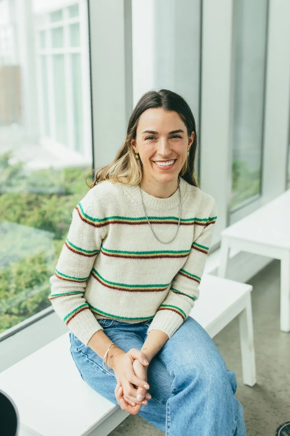 Catalina, a woman in a striped sweater and jeans, sits in front of a window and smiles into the camera