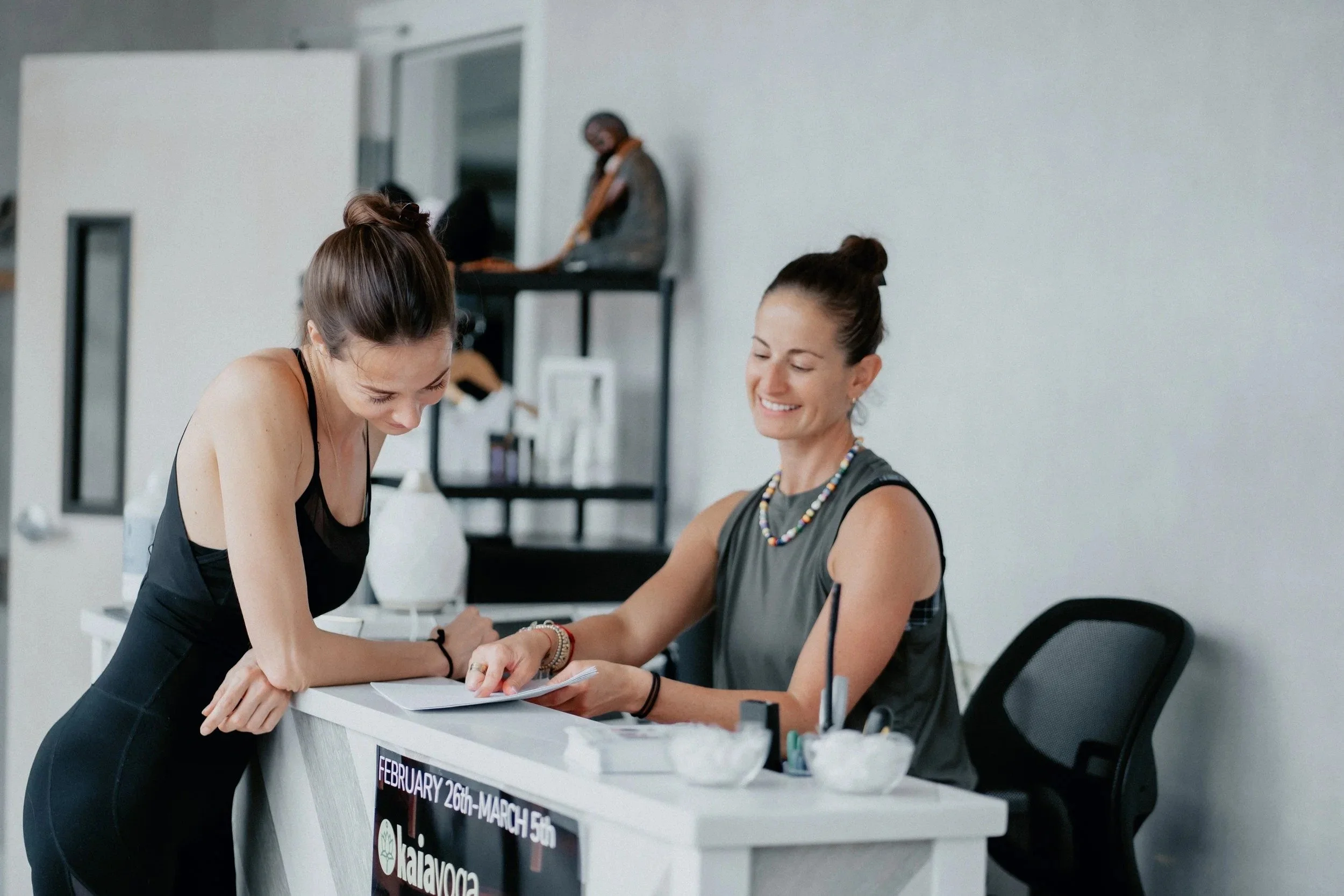 A woman in a black workout bodysuit looking at a page in a studio front desk as another woman explains something to her.