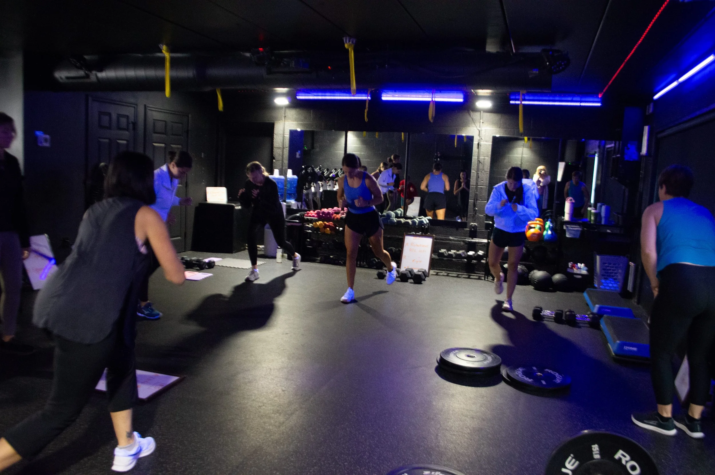 A dark-lighted gym room with blue neon lights where a group of women are doing strength training with dumbbells and weigth.