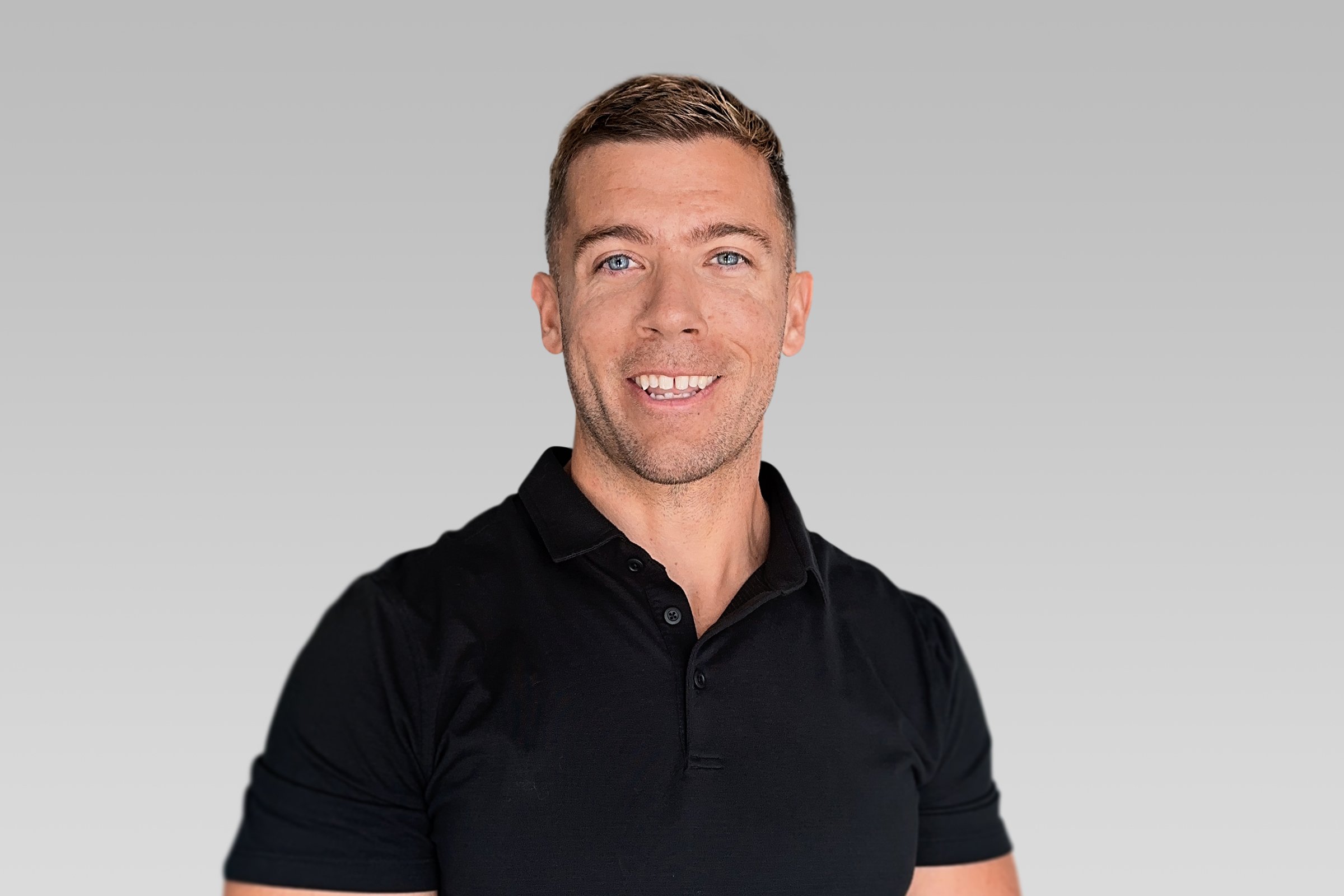 A smiling man with short brown hair and blue eyes wearing a black collared shirt, posing against a gray background.