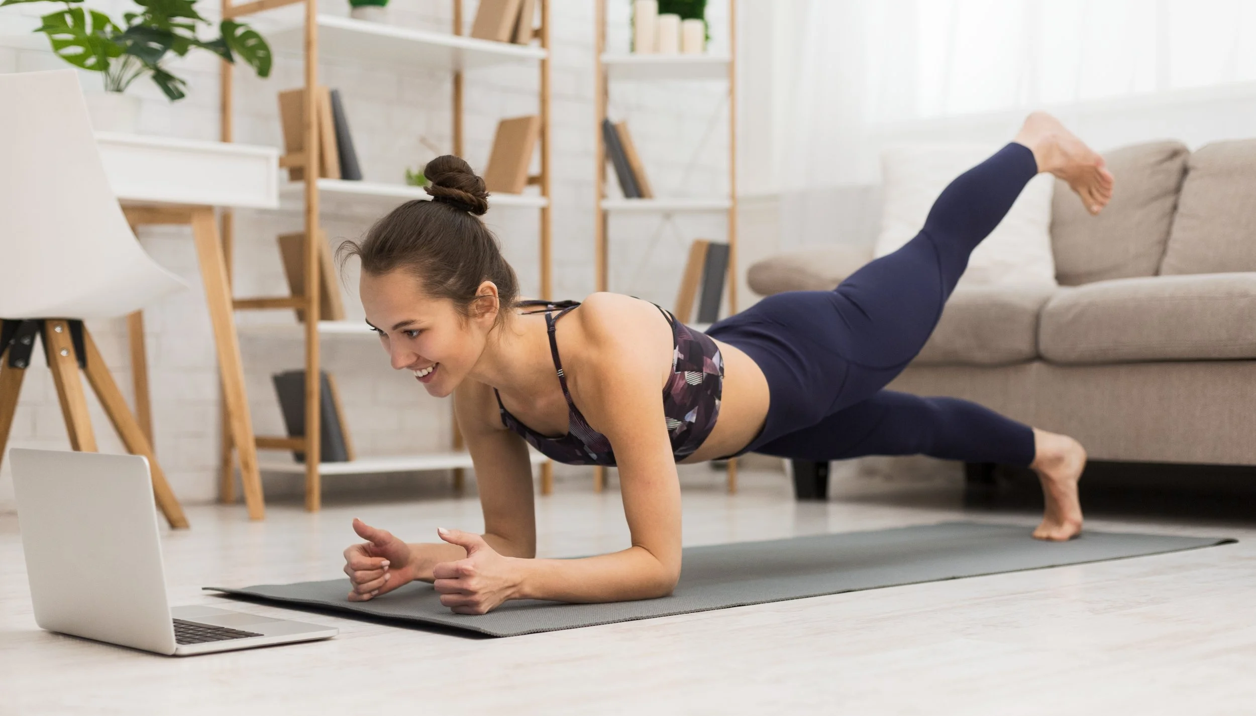 Woman doing mat Pilates in her living room with her laptop, during COVID-19 lockdown.