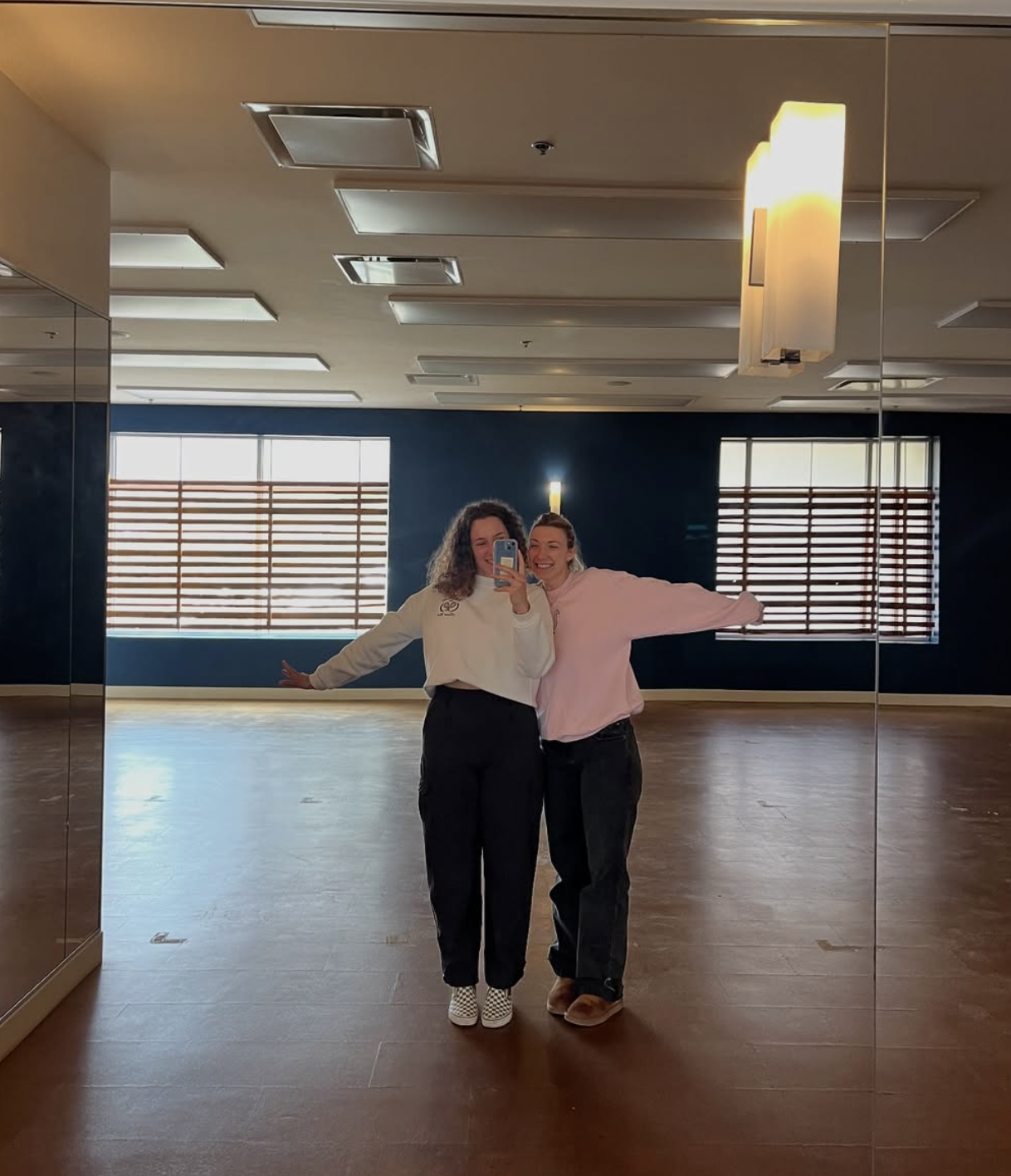 Two woman in sweat pants and sweaters, posing in front of a mirror in a boutique fitness studio room, while they take a selfie