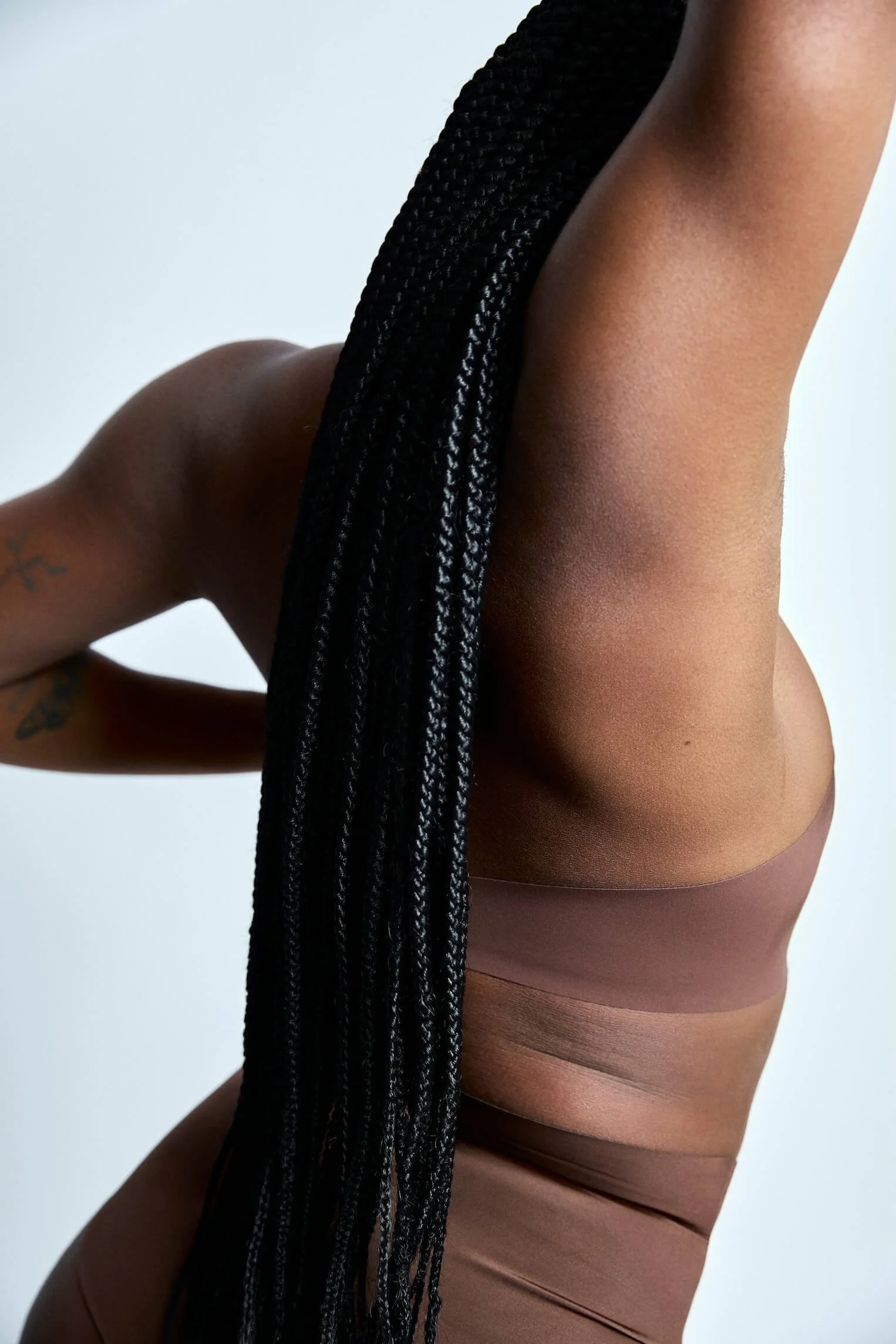 Close-up of a person with long black braided hair, wearing brown underwear, with a small tattoo on their arm, positioned against a plain light-colored background.
