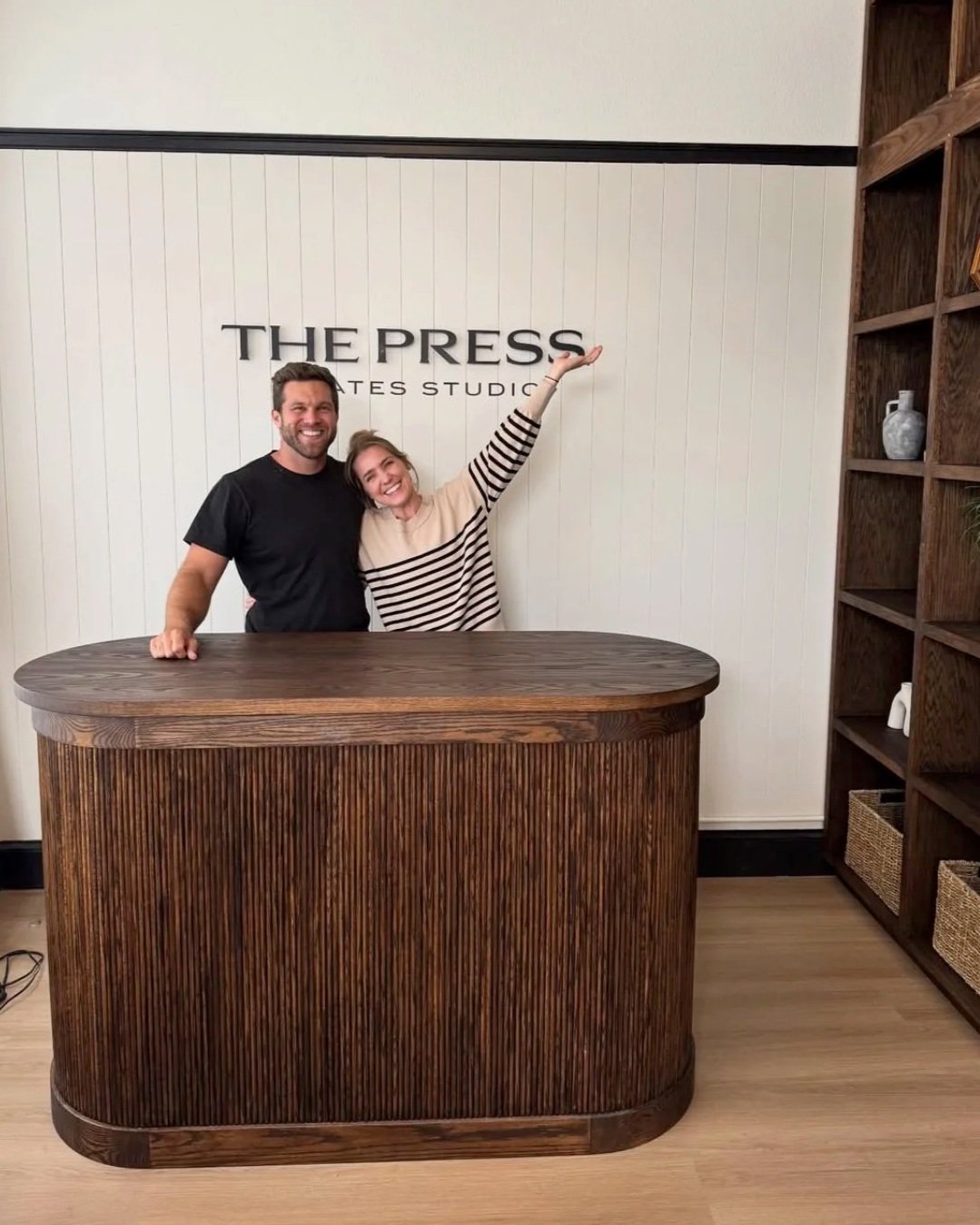 The Press Pilates owners standing behind the front desk as the woman points out with her hand to The Press Pilates sign on the wall.