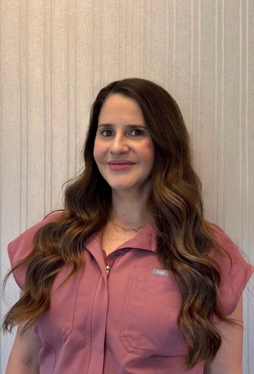 A woman with long wavy hair smiling, wearing a black scrub top with a name tag, standing in front of a white wall shelf with skincare products.
