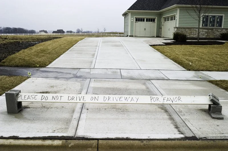 Freshly poured concrete driveway apron blocked by a wooden board handwritten in English and Spanish reading Please Do Not Drive on Driveway Por Favor, new ranch house with attached garage and horse fence behind, Chicago Illinois exurbs