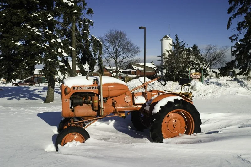 Vintage orange Allis-Chalmers tractor displayed as a lawn sculpture buried in snow in a small town center with a water tower, evergreen trees, and commercial buildings behind, Chicago Illinois exurbs