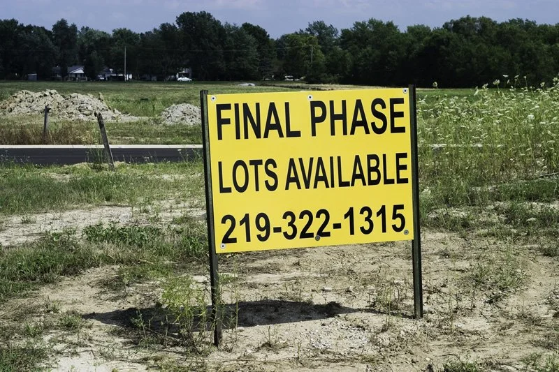 Yellow real estate sign reading Final Phase Lots Available with a phone number planted in a sandy vacant lot with weeds, a dirt road, gravel pile, and distant treeline in summer, Chicago Illinois exurbs