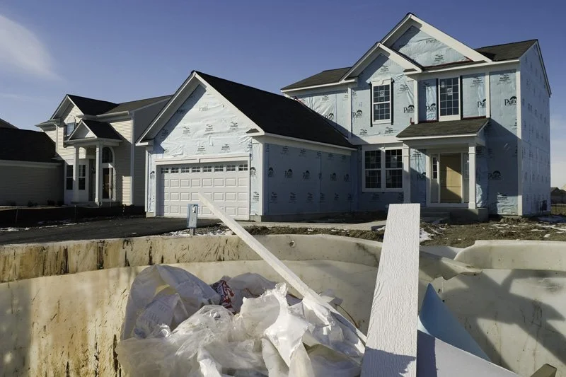 Two new suburban houses wrapped in blue housewrap with framed garage openings and unfinished facades, construction dumpster filled with trim material scraps in the foreground, frozen ground, Chicago Illinois exurbs