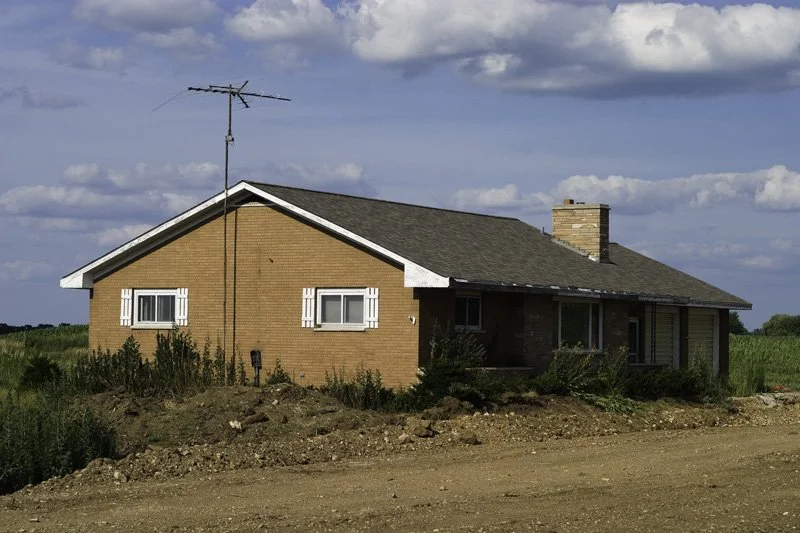 Single-story postwar brick ranch house with white shutters, chimney, and rooftop rabbit-ear TV antenna isolated on a dirt road beside a summer agricultural field under a cloudy sky, Chicago Illinois exurbs