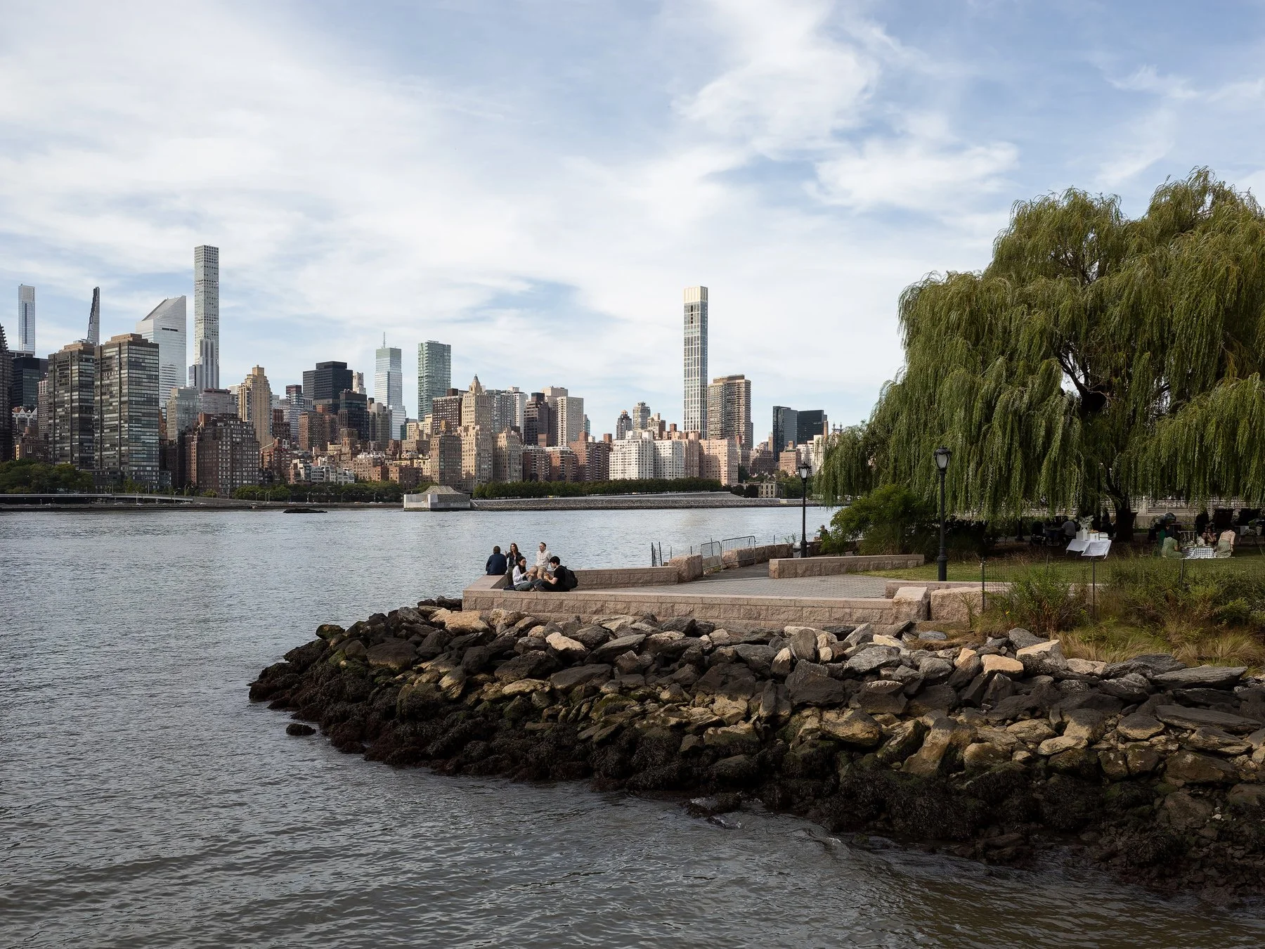 Sutton Tower in the Midtown Manhattan skyline seen across the East River from a riverfront park, with people resting near a willow tree and a rocky shoreline, by Thomas Juul-Hansen, New York New York - architectural photographer