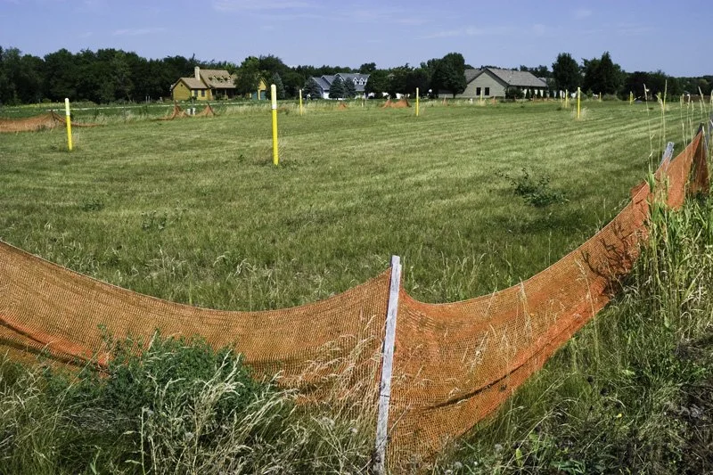 Stalled residential subdivision lot with sagging orange plastic silt fence, yellow survey stakes, overgrown grasses, and new houses visible on the horizon, Chicago Illinois exurbs