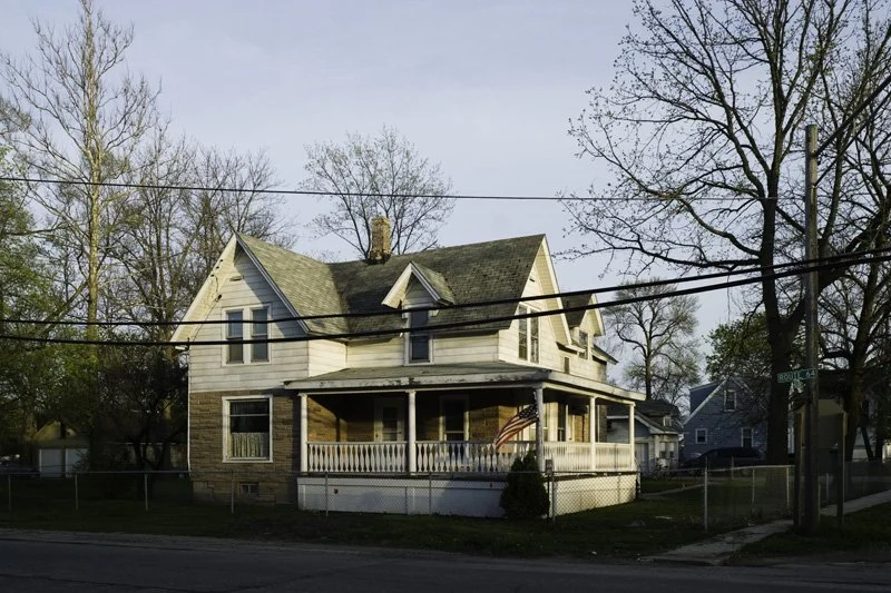 Aging two-story clapboard farmhouse with covered wraparound porch, American flag, chain link fence, and utility power lines on a corner lot surrounded by bare spring trees, Chicago Illinois exurbs