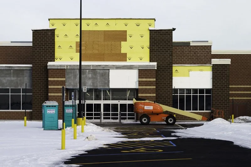 Big-box retail strip center under construction with yellow Tyvek weather barrier, orange boom lift, portable toilets, and snow-covered parking lot, Chicago Illinois exurbs