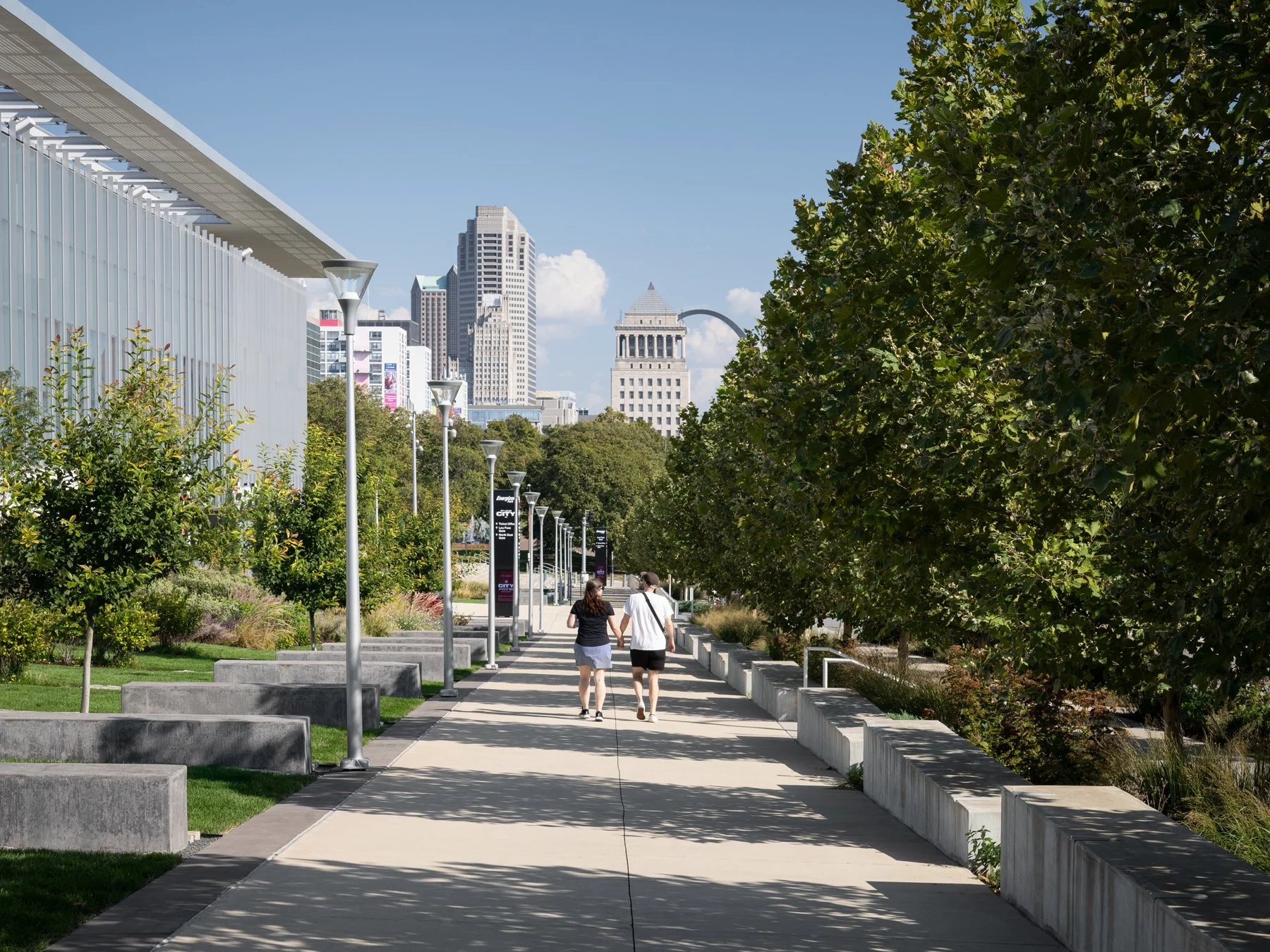 Energizer Park tree-lined promenade by day with a couple walking toward the downtown St. Louis skyline, Civil Courts Building, and Gateway Arch, landscape by DG2 Design, St. Louis Missouri - architectural photographer St. Louis Missouri