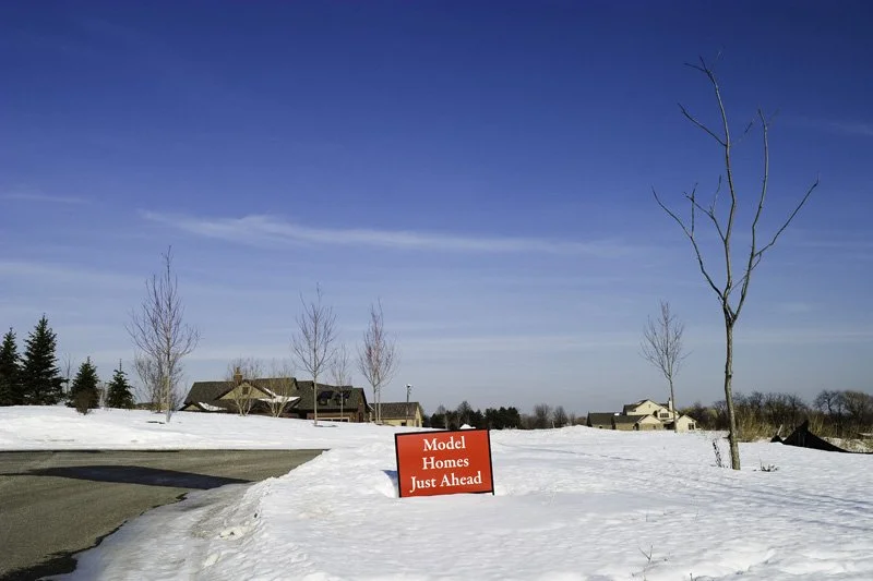 Red and white Model Homes Just Ahead real estate sign in a snow-covered field with bare nursery saplings, a lone deciduous tree, and new construction houses visible on the flat horizon under a blue sky, Chicago Illinois exurbs