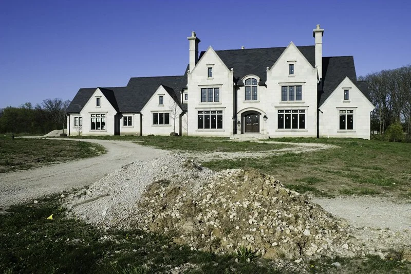 Large unfinished limestone Tudor Revival McMansion with multiple gabled wings, arched entry, and paired chimneys under construction, gravel and rubble pile in foreground on a spring day, Chicago Illinois exurbs