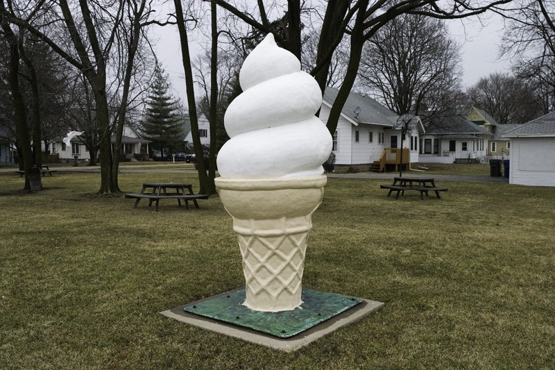 Oversized fiberglass soft-serve ice cream cone sculpture on a concrete base in a small town square park with wood picnic tables, bare deciduous trees, and modest residential houses behind, Chicago Illinois exurbs