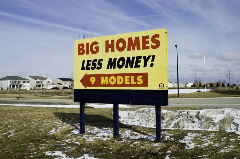 Large yellow freestanding billboard reading Big Homes Less Money 9 Models with an arrow, new subdivision houses and a streetlight visible behind patchy winter snow and dead grass, Chicago Illinois exurbs