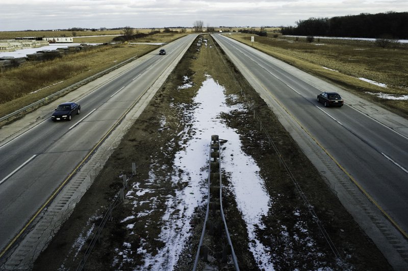 Elevated view looking down a divided four-lane highway with a snow-patched grassy median, railroad tracks below, flat agricultural land, sparse commercial development, and two cars receding to the horizon, Chicago Illinois exurbs