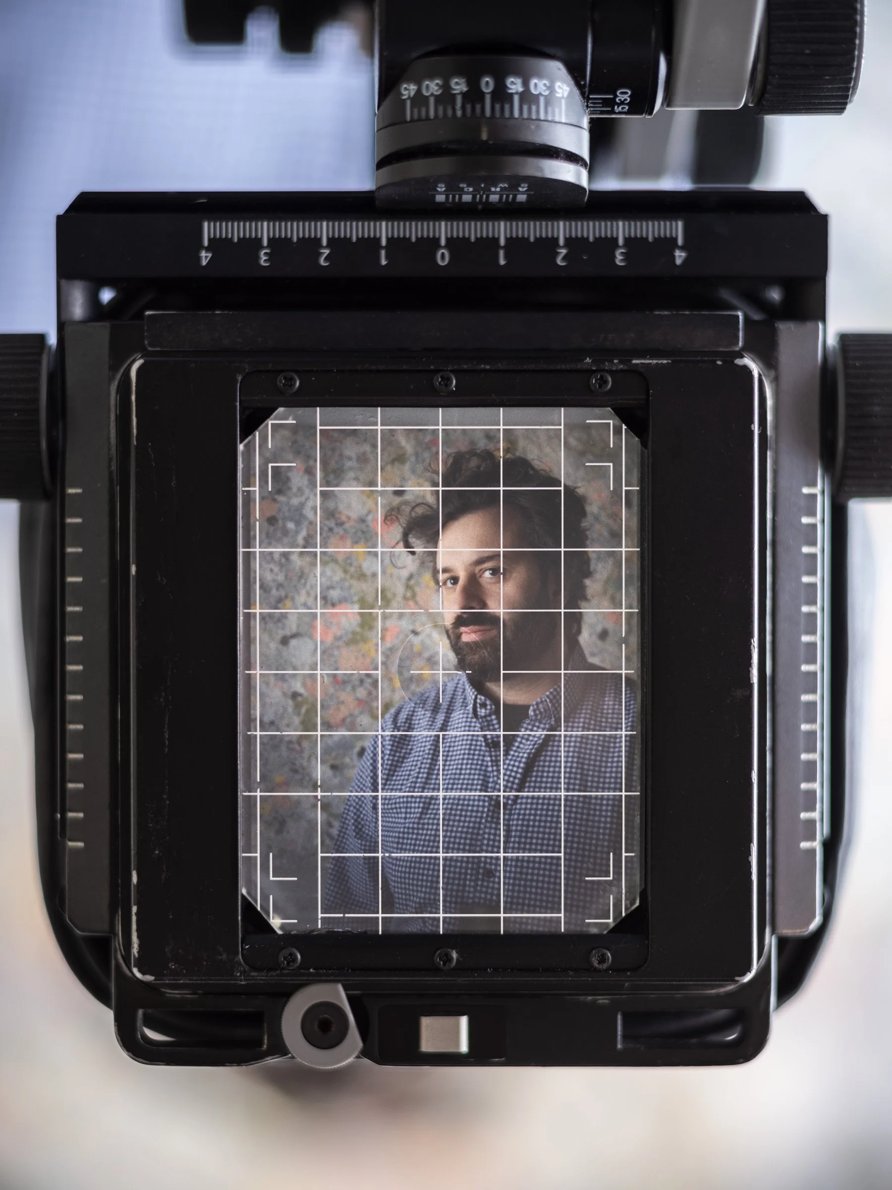 Camera viewfinder showing a portrait of a man with dark curly hair and beard, wearing a checkered shirt, in front of a colorful textured background.