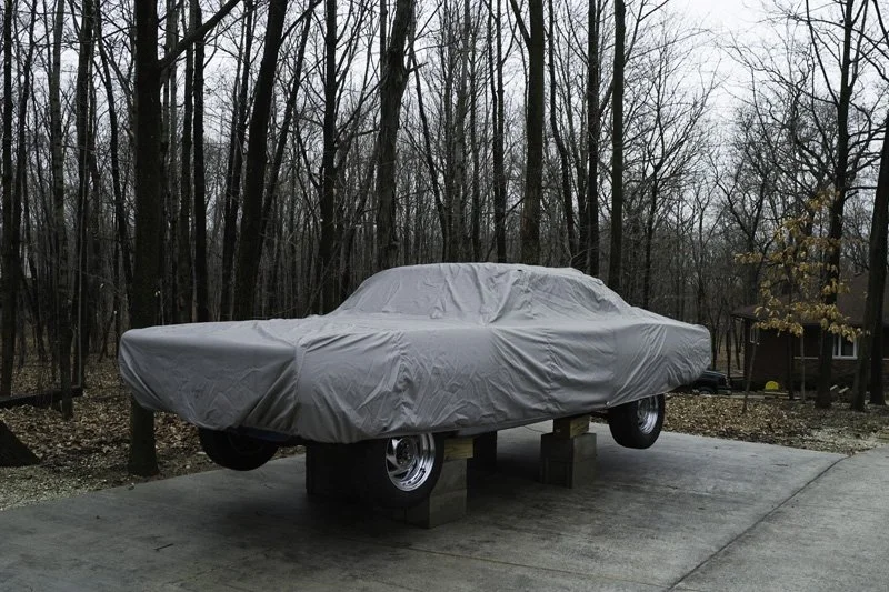 Vintage American muscle car stored on wooden jack stands under a gray fitted cover on a concrete driveway surrounded by bare winter deciduous trees and a brick house, Chicago Illinois exurbs