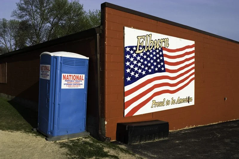 Painted American flag mural reading Elburn Proud to be American on the exterior wall of a red brick commercial building beside a blue portable toilet in morning light, Chicago Illinois exurbs