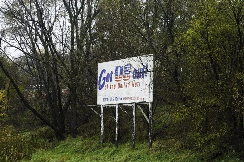 Weathered roadside billboard on white-painted wooden posts reading Get US Out of the United Nations in patriotic lettering surrounded by overgrown autumn trees and shrubs, Chicago Illinois exurbs