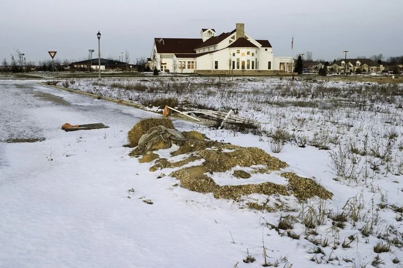Abandoned snowy residential development site with gravel and debris pile, collapsed construction materials, bare scrub vegetation, and a white gabled community building with a flag in the background, Chicago Illinois exurbs