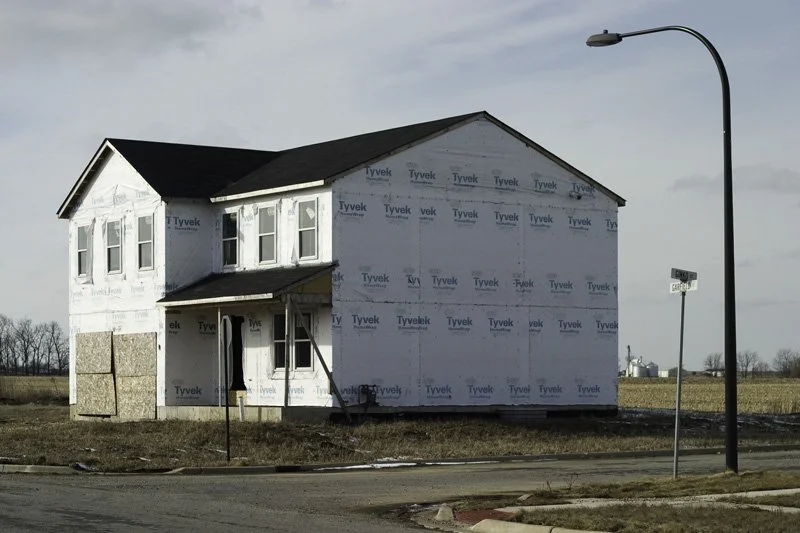 Unfinished two-story house fully wrapped in white Tyvek housewrap on a bare corner lot beside a cornfield and a single street lamp under a gray winter sky, Chicago Illinois exurbs