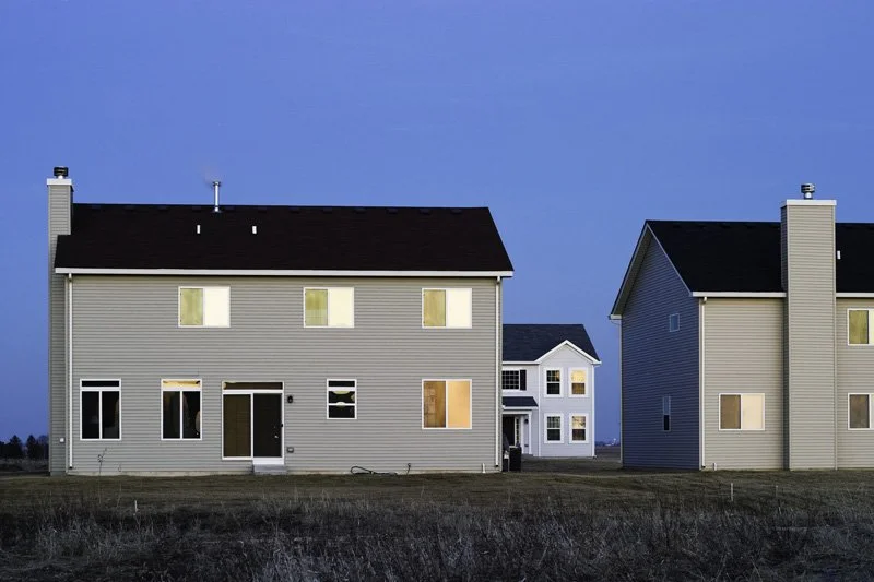 Three vinyl-sided new construction houses with illuminated windows at blue hour facing an open grass field under a deep twilight sky, Chicago Illinois exurbs