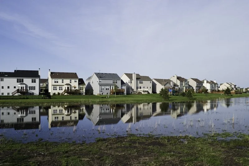 Row of new two-story suburban tract houses with vinyl siding and attached garages reflected in a calm retention pond under a wide open sky, Chicago Illinois exurbs