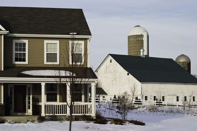 New craftsman-style two-story house with front porch beside an old white painted barn, concrete grain silos, and split rail fence in winter sunlight against a pale blue sky, Chicago Illinois exurbs