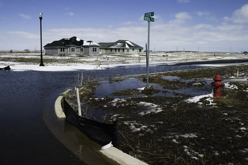 Flooded new residential street intersection with submerged curb, street signs reading Aspen and Deerfield, red fire hydrant, erosion barrier, and a lone single-story house on a flat open snowy plain, Chicago Illinois exurbs