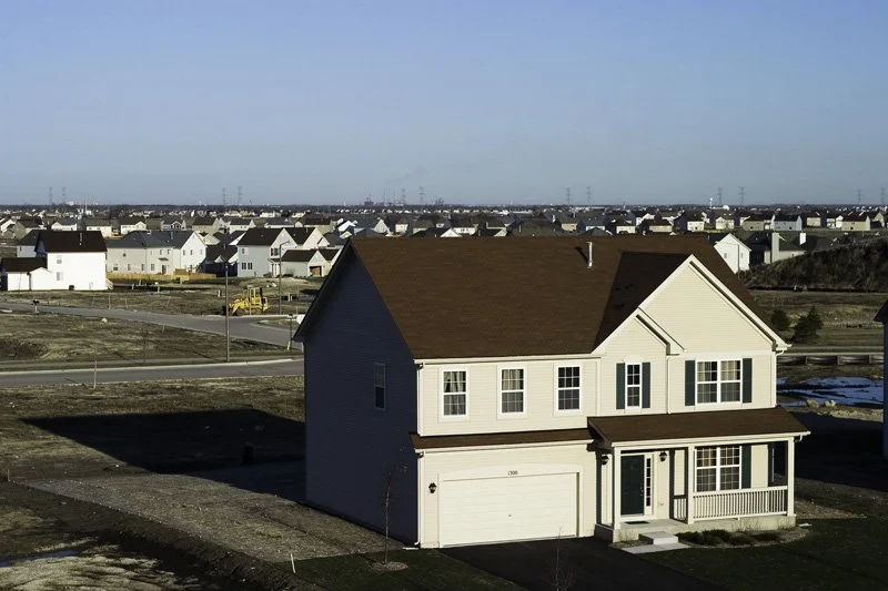 Elevated view of a new two-story vinyl-sided model home in the foreground with a sprawling tract housing subdivision, bare graded lots, construction equipment, and high-voltage power line towers receding to the flat horizon, Chicago Illinois exurbs