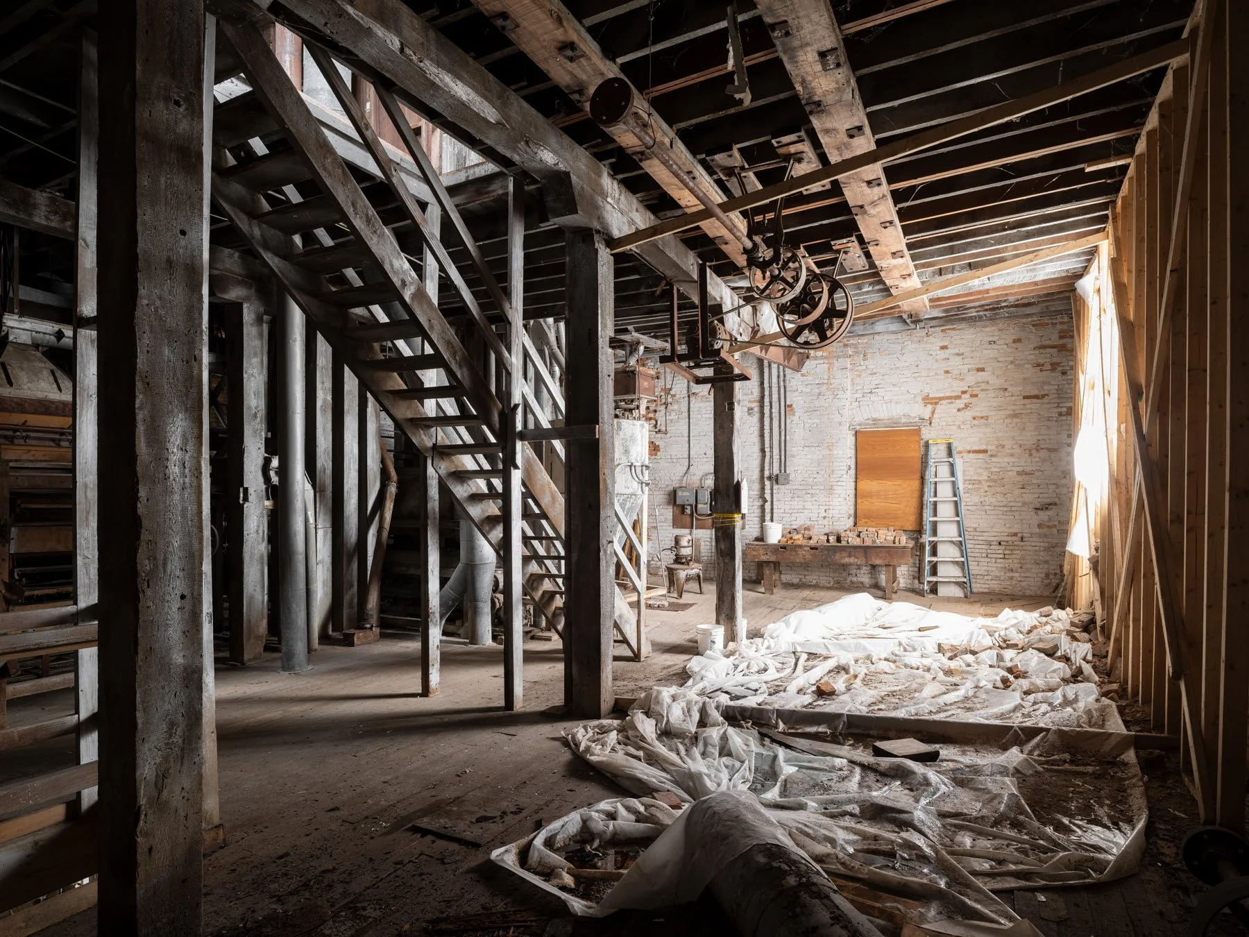 Millstadt Mill interior with timber staircase, overhead line-shaft pulley, and repair dropcloths on the floor - architectural photographer St. Louis Missouri