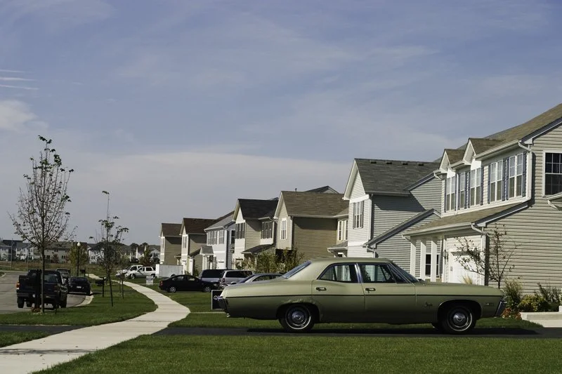 New residential subdivision street with a vintage green Chevrolet Impala sedan parked at the curb, young nursery-planted street trees, and attached garage townhomes receding into the distance, Chicago Illinois exurbs