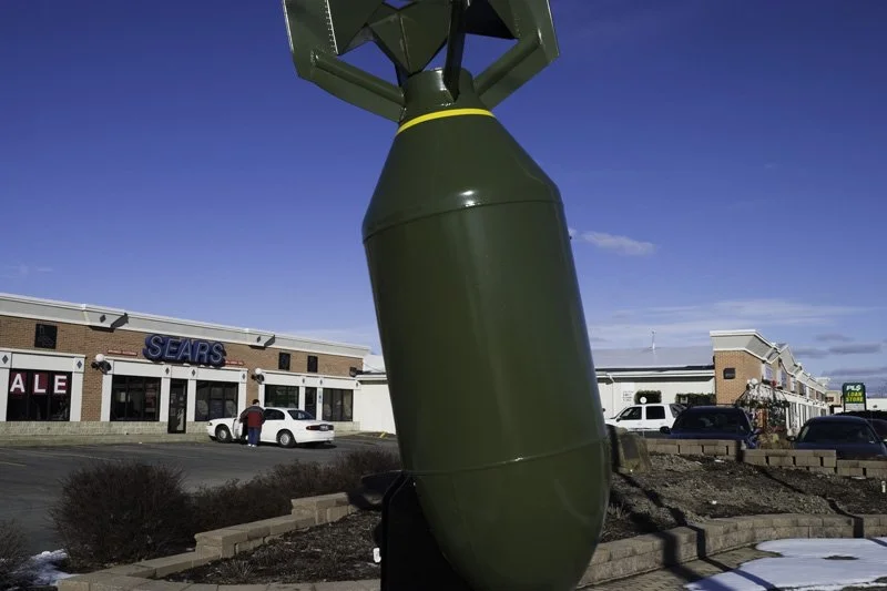 Oversized painted military bomb sculpture on a masonry planter base in a strip mall parking lot with a Sears storefront, sale signage, and parked cars visible behind under a blue winter sky, Chicago Illinois exurbs