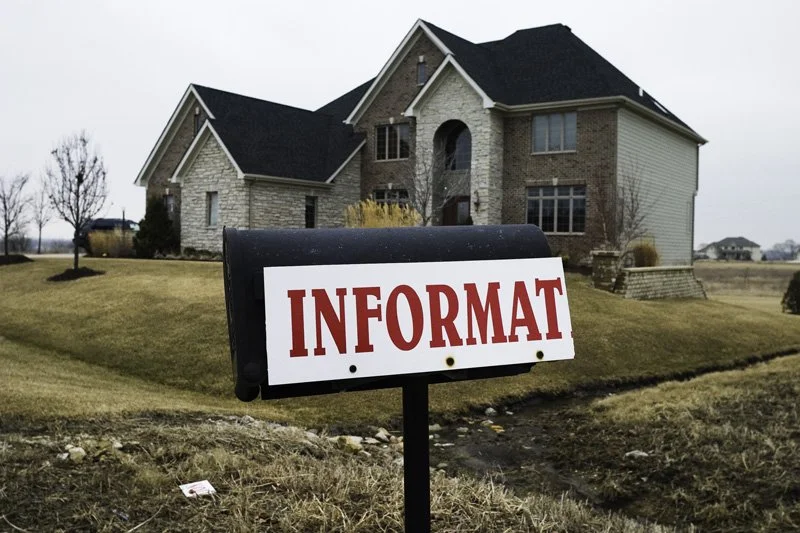 Black mailbox post with a truncated real estate information sign reading INFORMAT in front of a two-story brick and stone McMansion with arched entry and attached garage on a gray winter day, Chicago Illinois exurbs