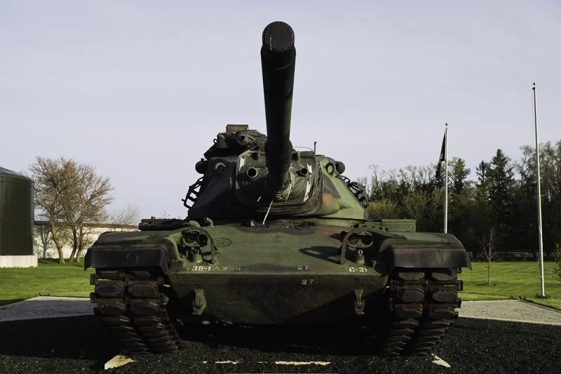 Decommissioned camouflage military tank with long gun barrel displayed on a paved pad in front of a flagpole and manicured lawn beside a commercial building, Chicago Illinois exurbs