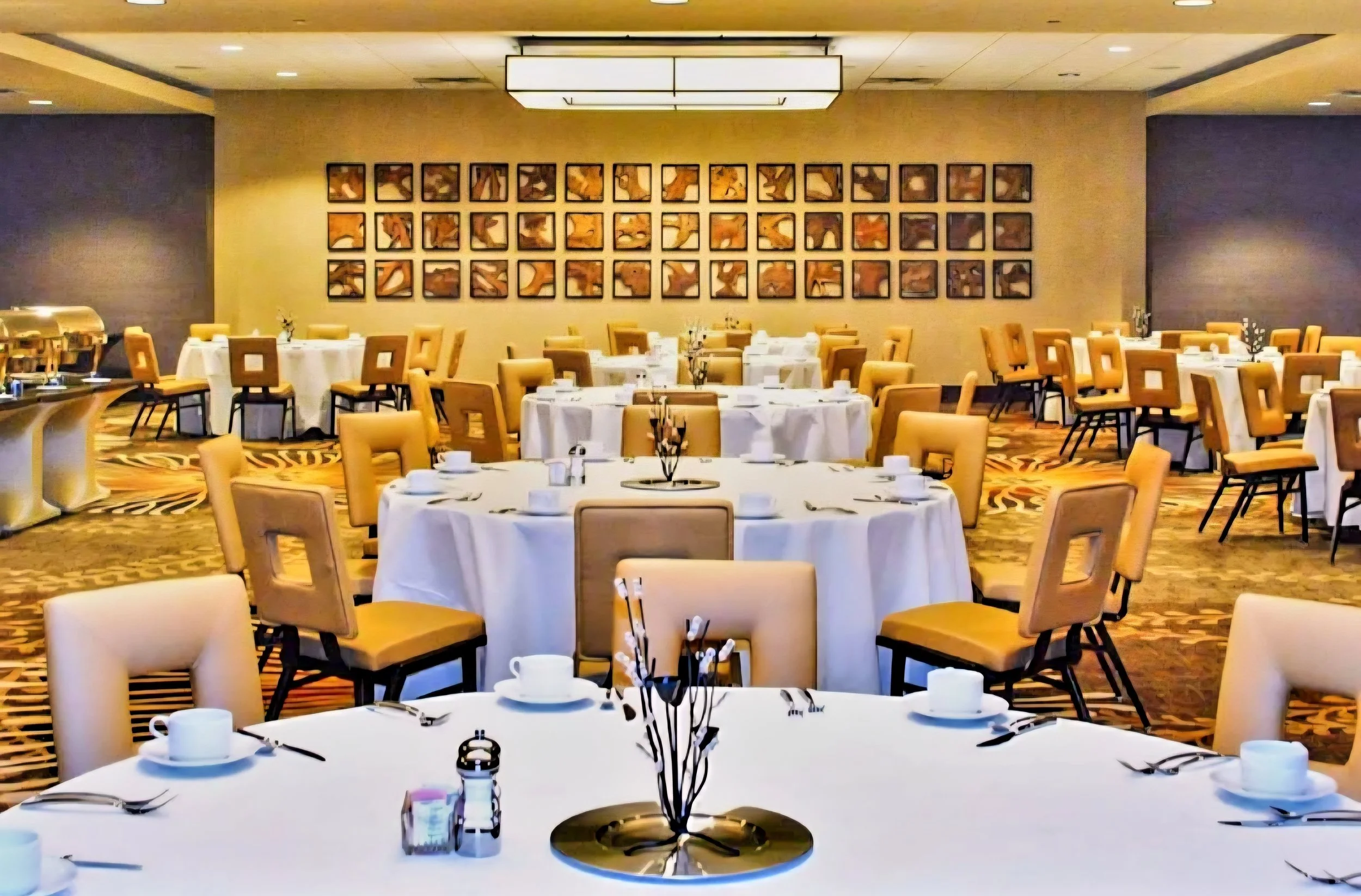 Dining room with round tables set with white tablecloths, cups, and silverware. A wall featuring a grid of framed abstract artwork. Yellow upholstered chairs around the tables and patterned carpet flooring.