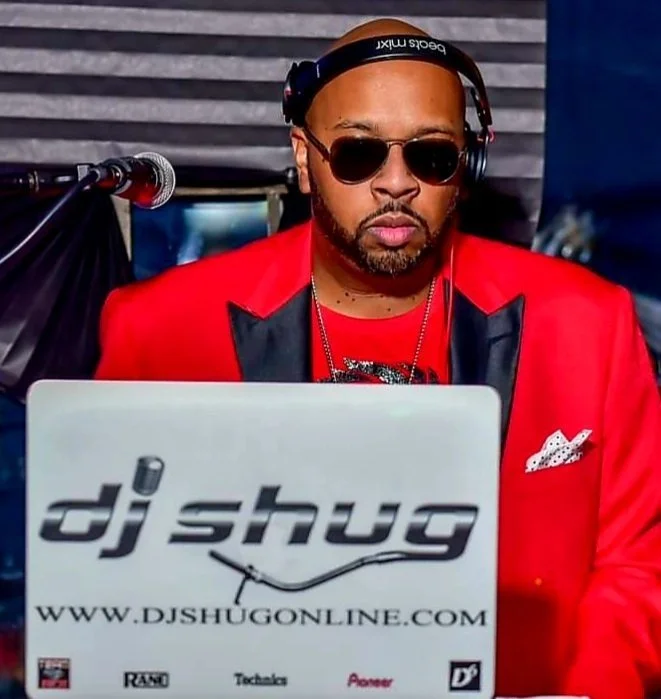 Male DJ wearing sunglasses, headphones, and a red blazer, working at a DJ booth with a laptop displaying 'dj shug' and website.