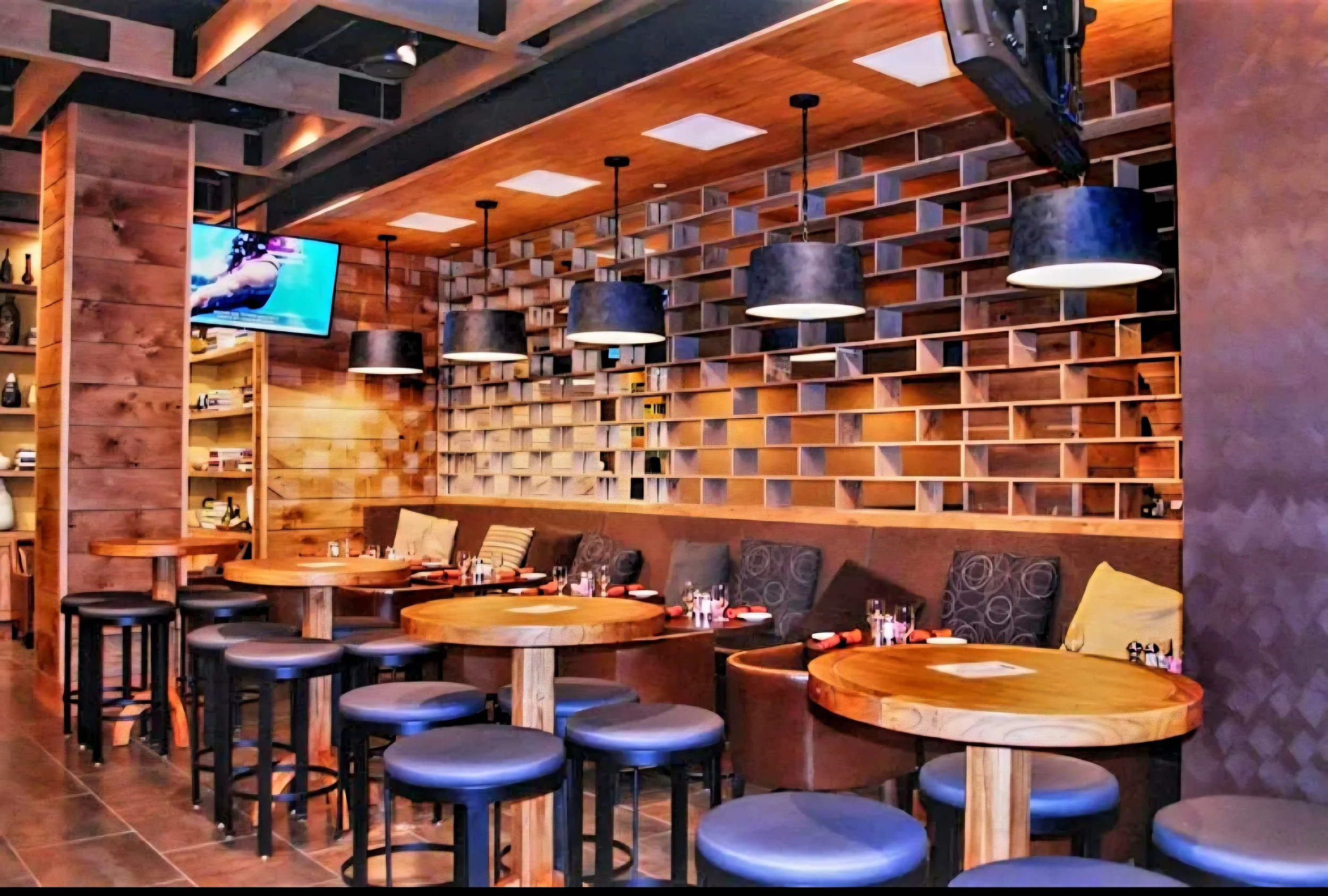 Interior of a restaurant with round wooden tables, blue cushioned stools, a wooden wall with a geometric pattern, a TV mounted on the wall, and hanging black pendant lights.