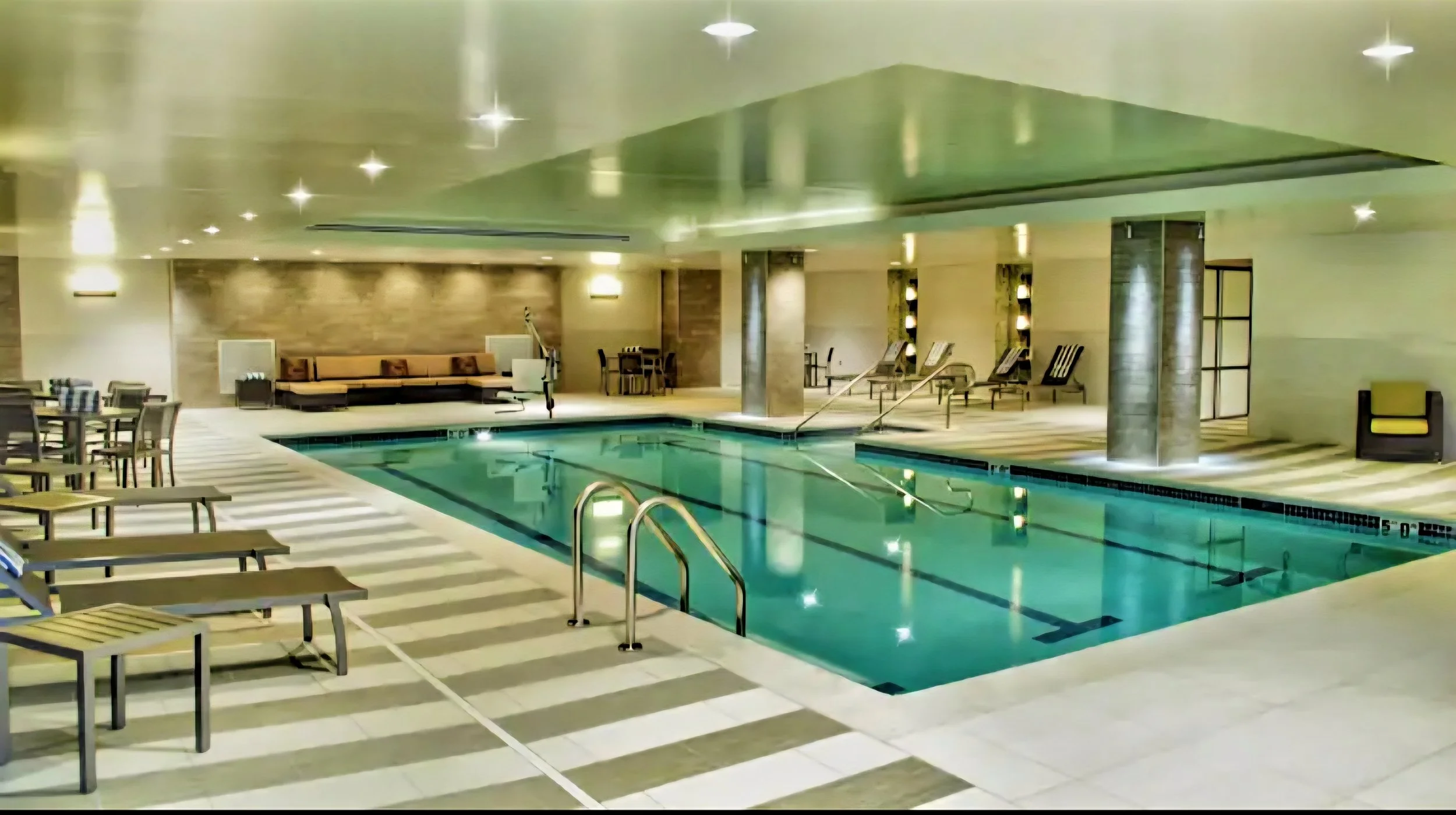 Indoor hotel swimming pool with lounge chairs, tables, and a seating area, illuminated by soft lighting and a reflective ceiling.