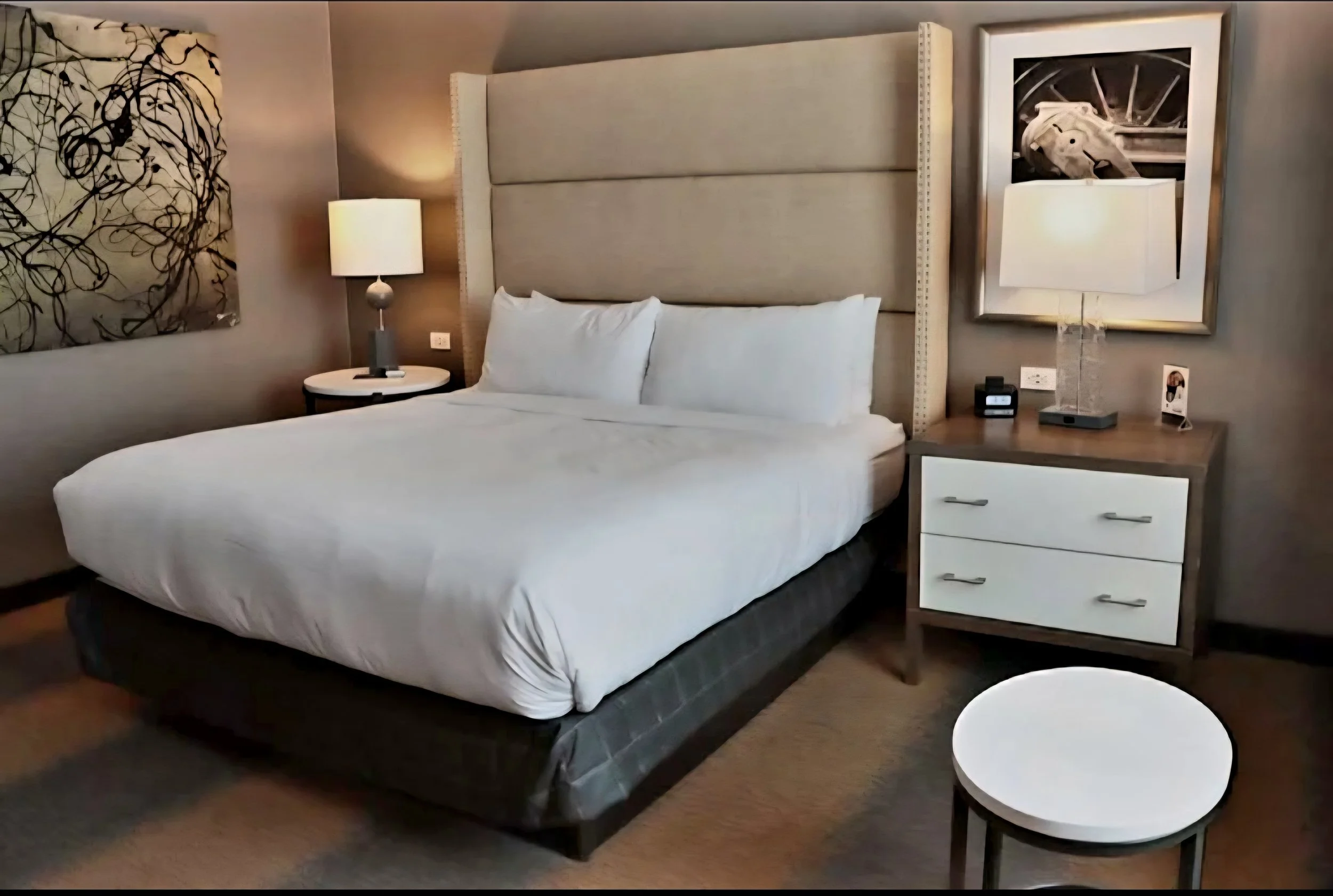 A hotel room with a bed, nightstands, lamps, abstract artwork, a framed horse print, and a small round table.