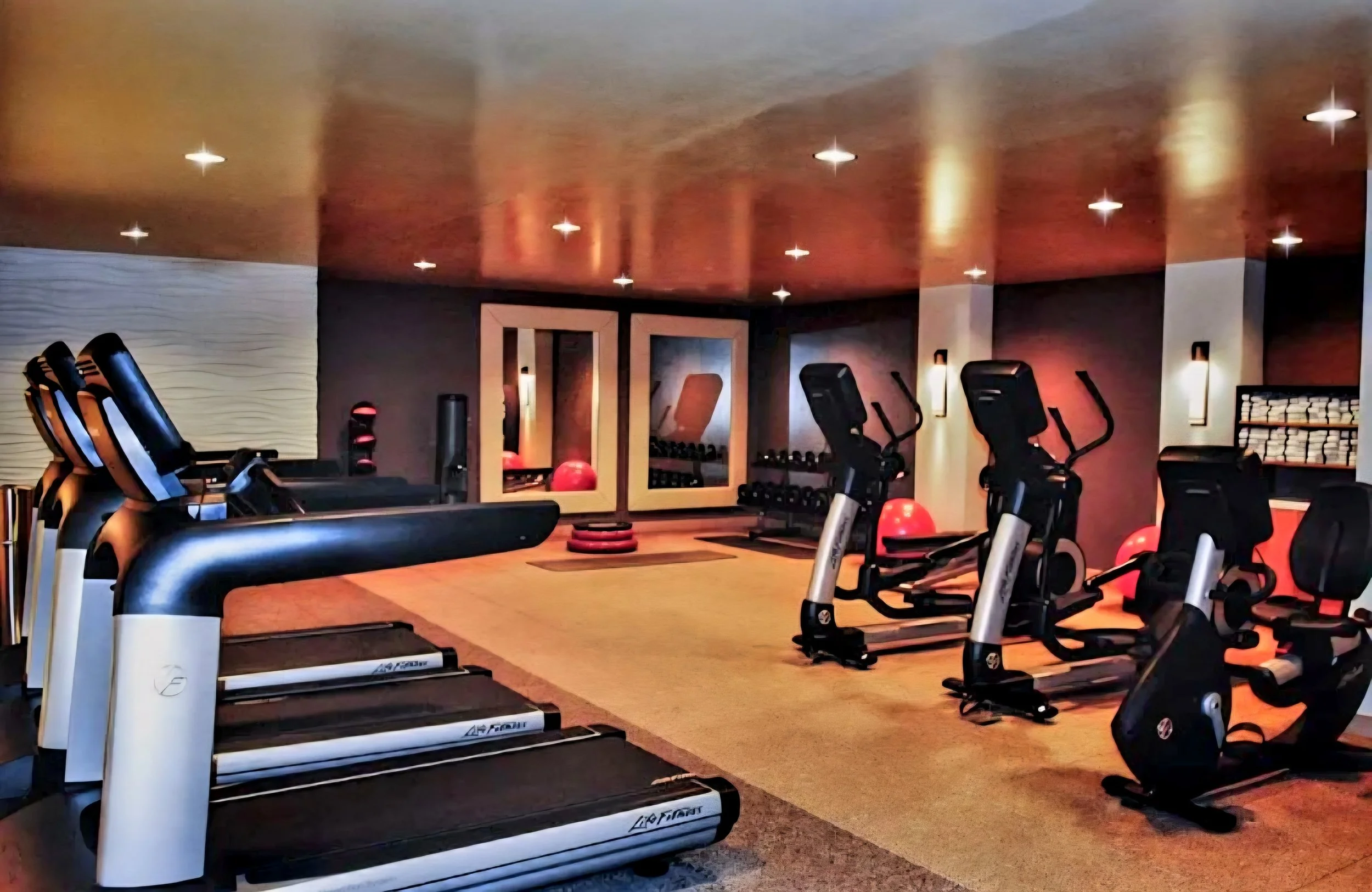 A fitness gym with treadmills, exercise bikes, medicine balls, and weight racks, featuring warm lighting and modern decor.